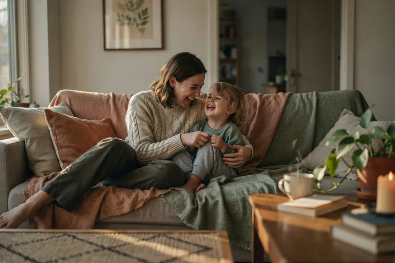 A parent and child cuddled under terracotta and sage throw blankets on a cozy sofa in a warmly lit Canadian living room